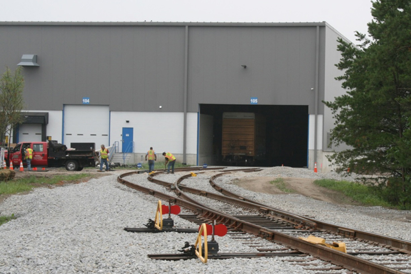FIGURE 3. Two-Track Siding Inside Expanded Warehouse
This image shows two railroad tracks entering a modern warehouse. There is a large boxcar positioned on one track for indoor loading and unloading.
FIGURE 3. Two-Track Siding Inside Expanded Warehouse
This image shows two railroad tracks entering a modern warehouse. There is a large boxcar positioned on one track for indoor loading and unloading.